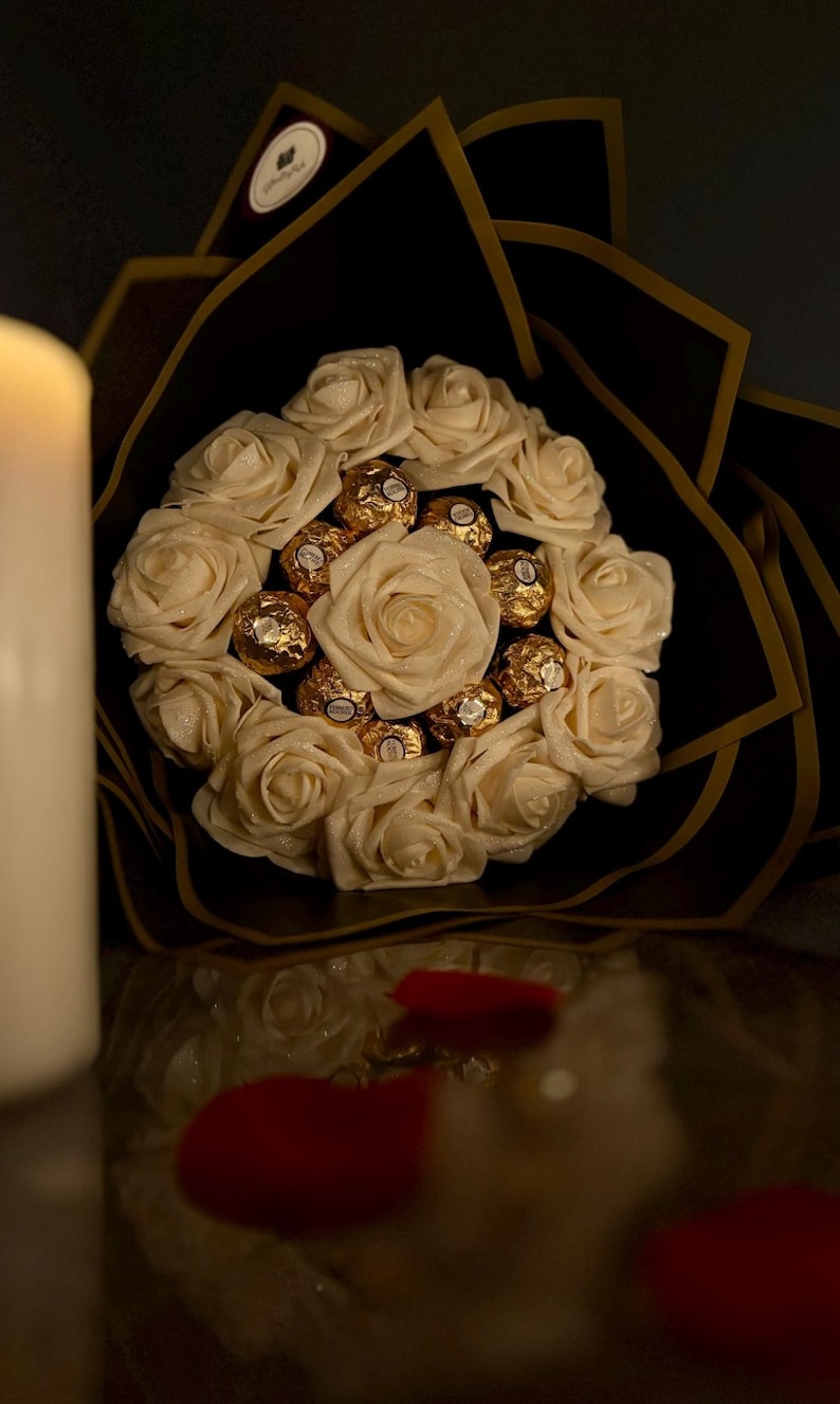 May include: A bouquet of cream-colored roses and gold-wrapped chocolates. The arrangement is set against a black background with gold accents. Red rose petals are scattered near the bouquet. A white candle is in the background.