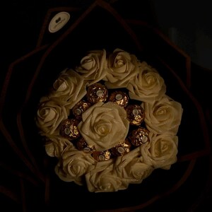 May include: A bouquet of cream-colored roses with gold-wrapped chocolate candies nestled among the petals. The arrangement is set against a dark background, creating a striking contrast. The roses appear to have a subtle shimmer.