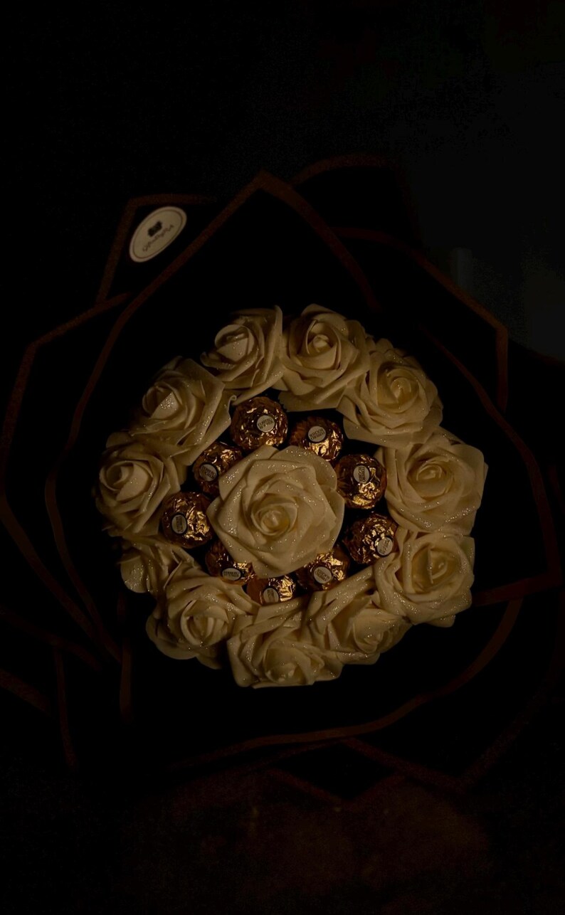May include: A bouquet of cream-colored roses with gold-wrapped chocolate candies nestled among the petals. The arrangement is set against a dark background, creating a striking contrast. The roses appear to have a subtle shimmer.