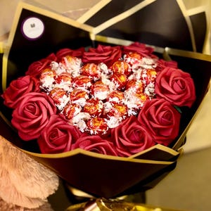 May include: A bouquet of red roses and Lindor chocolate candies, wrapped in black and gold paper. The bouquet is tied with a gold ribbon and is presented on a neutral background.