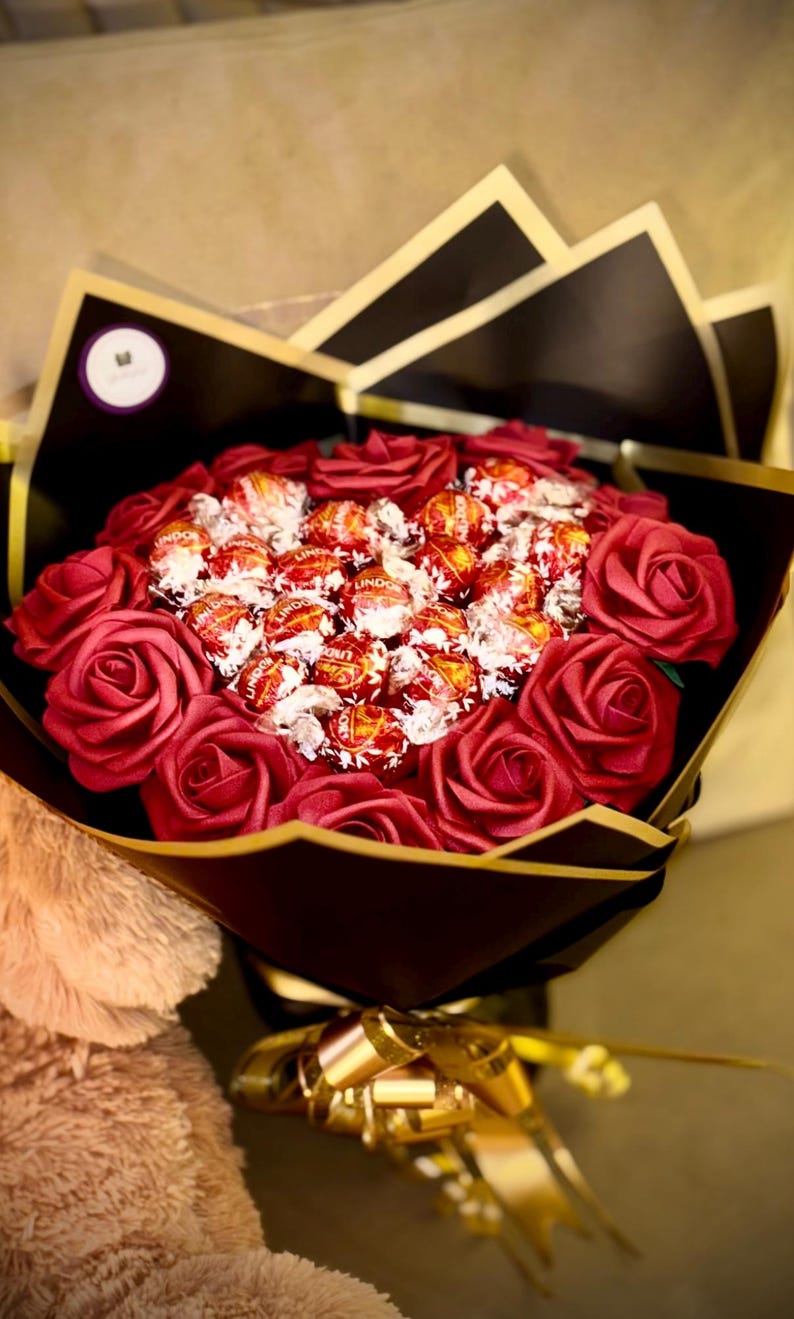 May include: A bouquet of red roses and Lindor chocolate candies, wrapped in black and gold paper. The bouquet is tied with a gold ribbon and is presented on a neutral background.