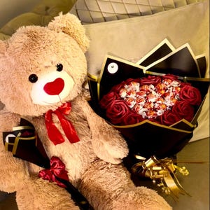May include: A large, plush teddy bear with a red nose and bow, next to a bouquet of red roses and wrapped candies. The bear has red heart-shaped accents on its feet. The bouquet is wrapped in black and gold paper.