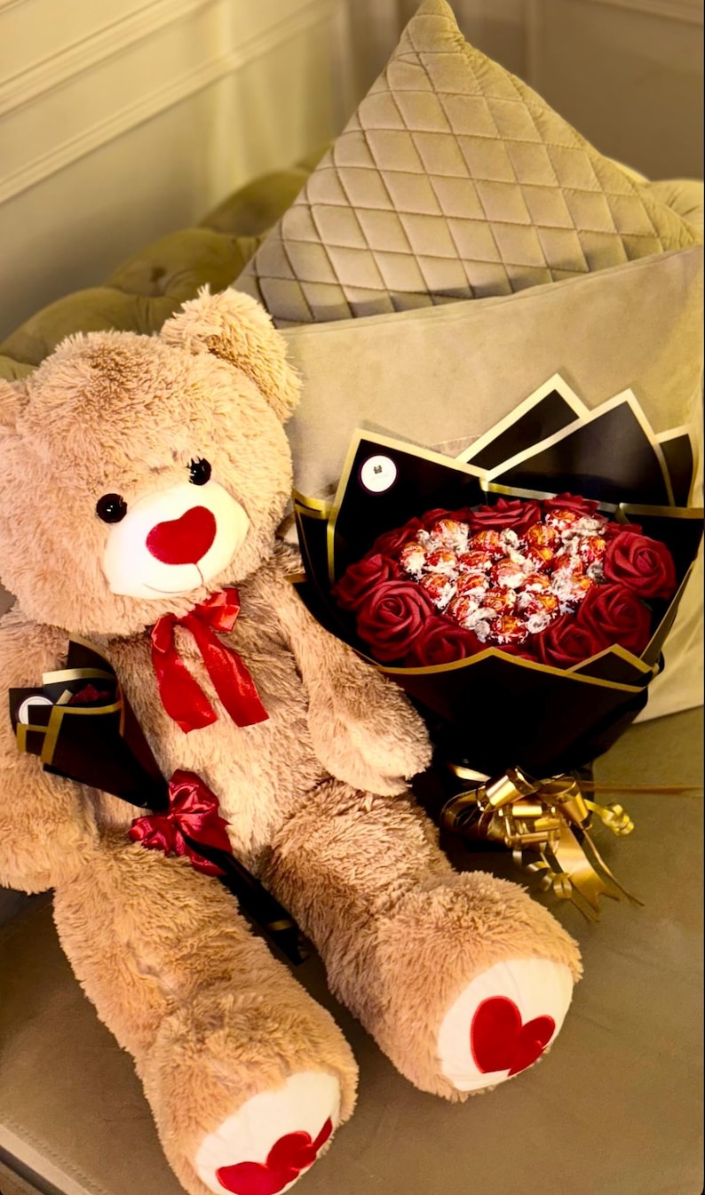May include: A large, plush teddy bear with a red nose and bow, next to a bouquet of red roses and wrapped candies. The bear has red heart-shaped accents on its feet. The bouquet is wrapped in black and gold paper.
