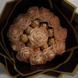 May include: A bouquet of light pink roses and gold-wrapped chocolates, arranged in a circular pattern. The bouquet is wrapped in black and gold paper, creating a visually appealing gift.