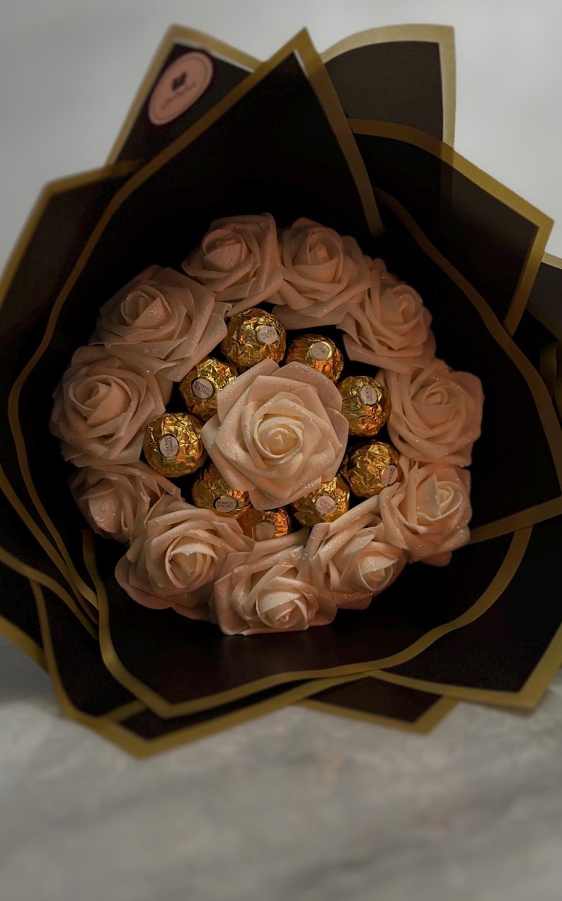 May include: A bouquet of light pink roses and gold-wrapped chocolates, arranged in a circular pattern. The bouquet is wrapped in black and gold paper, creating a visually appealing gift.