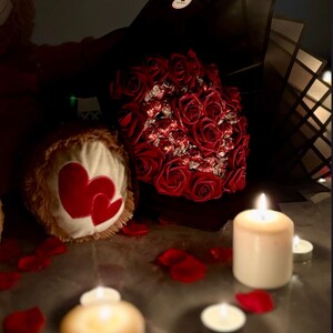 May include: A romantic scene featuring a bouquet of red roses, a teddy bear with heart-shaped accents, scattered rose petals, and lit candles. The soft lighting creates a warm and intimate atmosphere.
