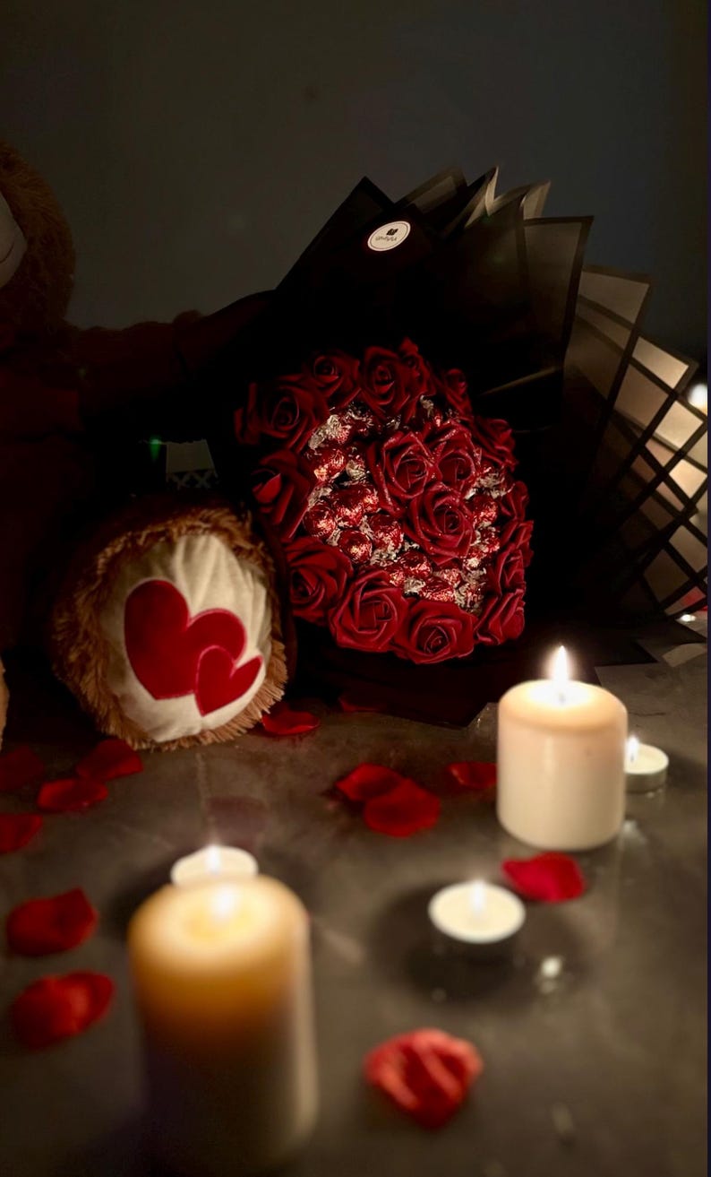 May include: A romantic scene featuring a bouquet of red roses, a teddy bear with heart-shaped accents, scattered rose petals, and lit candles. The soft lighting creates a warm and intimate atmosphere.