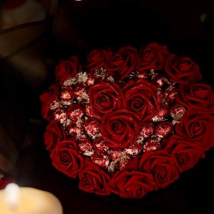 May include: A heart-shaped arrangement of red roses and chocolate candies. The roses are arranged in a heart shape, with the candies filling the center. The bouquet is wrapped in dark paper, creating a romantic and elegant presentation.