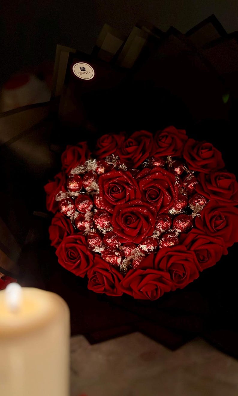 May include: A heart-shaped arrangement of red roses and chocolate candies. The roses are arranged in a heart shape, with the candies filling the center. The bouquet is wrapped in dark paper, creating a romantic and elegant presentation.