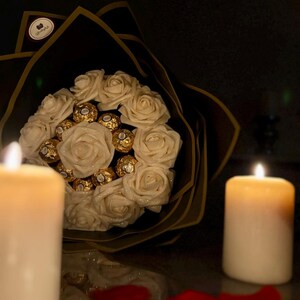 May include: A bouquet of white roses and gold-wrapped chocolates, wrapped in black paper with gold trim. The arrangement is set against a dark background, with lit candles and scattered red rose petals.