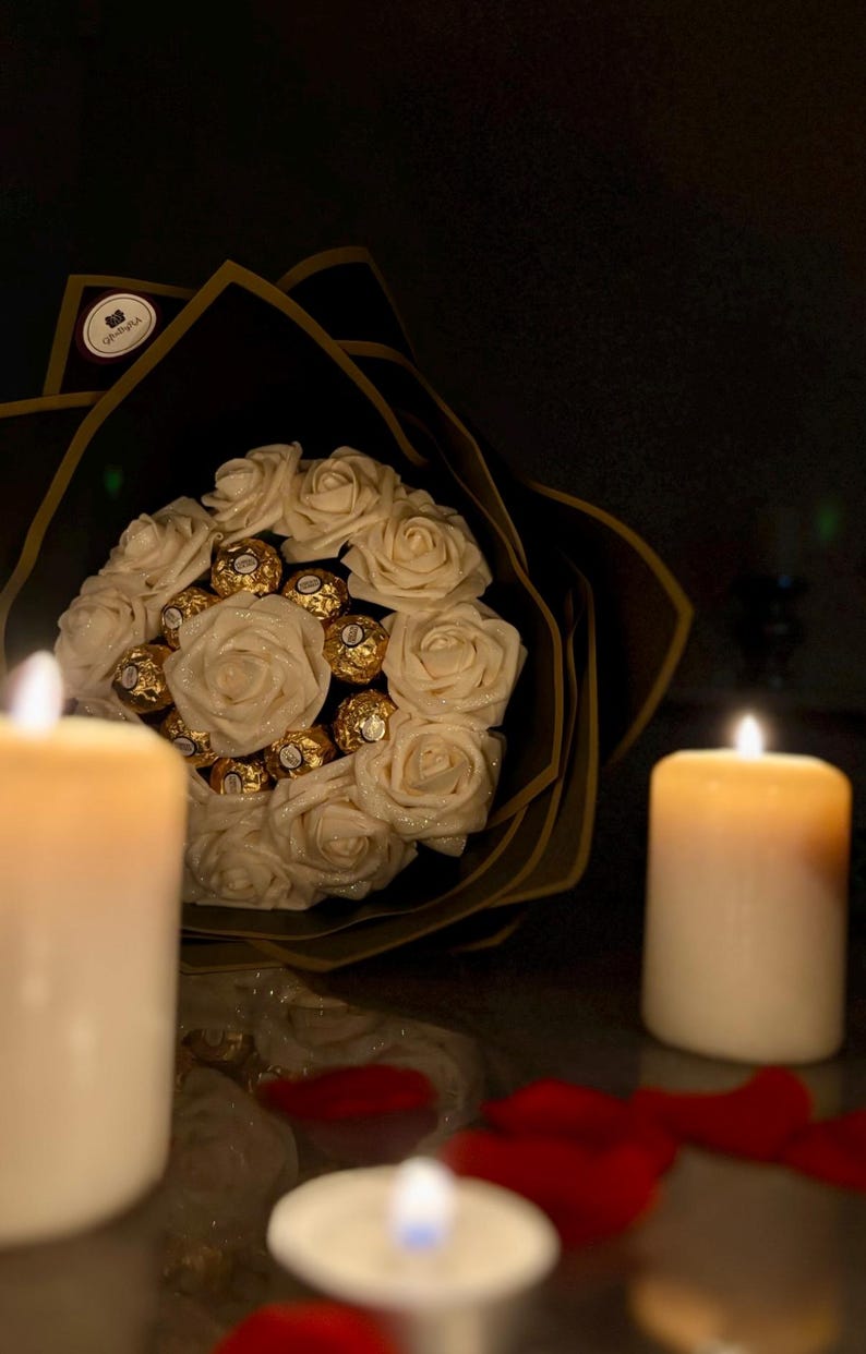 May include: A bouquet of white roses and gold-wrapped chocolates, wrapped in black paper with gold trim. The arrangement is set against a dark background, with lit candles and scattered red rose petals.