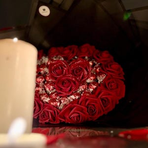 May include: A bouquet of red roses, artfully arranged with chocolates, is presented against a dark backdrop. A lit candle in the foreground adds a warm glow to the romantic floral arrangement.