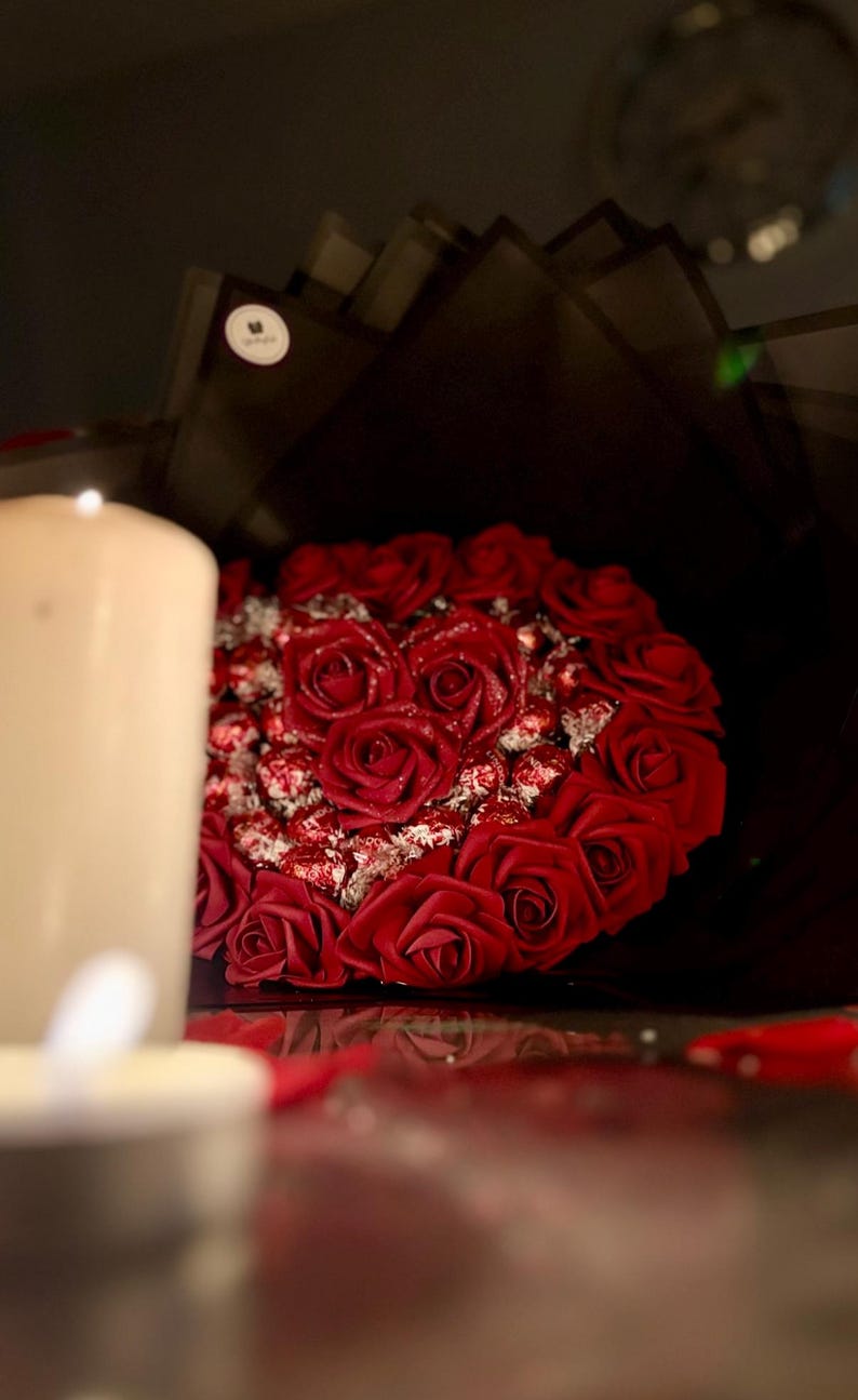 May include: A bouquet of red roses, artfully arranged with chocolates, is presented against a dark backdrop. A lit candle in the foreground adds a warm glow to the romantic floral arrangement.