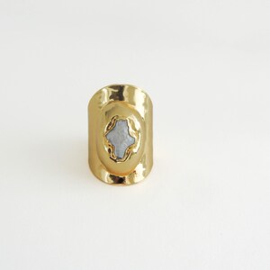 May include: A gold-colored ring with an oval shape and a light blue stone in the center. The ring has a smooth, polished finish and a wide band. The stone has a unique, irregular shape, adding to the ring's distinctive design.