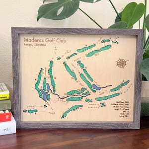 May include: Framed wooden map of Moderas Golf Club in Poway, California, with green and blue course details. The frame is gray. A white and green lamp sits on a stack of books, and a green figurine is on the right.