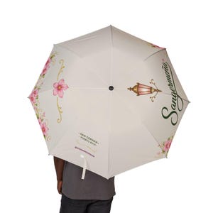 May include: A white umbrella with a floral design and the text "Sangermeña" in green. The umbrella features pink flowers and a decorative lamp illustration. Additional text includes "San German - Puerto Rico" and a social media handle.