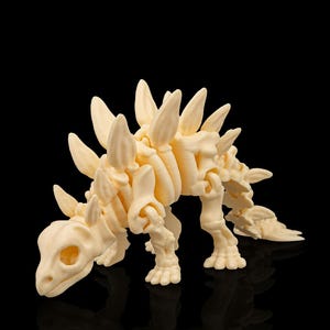 Può includere: Statuetta di scheletro di dinosauro Stegosaurus color crema. Il modello ha un design dettagliato, comprese le iconiche placche lungo la schiena e un cranio dettagliato. La statuetta è ambientata su uno sfondo nero.