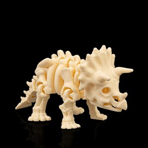 Puede incluir: Modelo de esqueleto de dinosaurio Triceratops de color crema. El modelo tiene una estructura esquelética detallada, incluyendo el cráneo con cuernos, volante y patas articuladas. El dinosaurio está sobre un fondo negro.