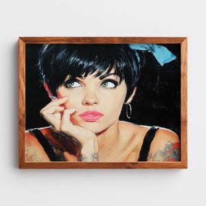 Puede incluir: Retrato enmarcado de una mujer con pelo corto negro, ojos verdes y lápiz labial rosa. Lleva un top negro y tiene tatuajes en el brazo. Un lazo azul claro está en su pelo. La obra de arte está en un marco de madera.