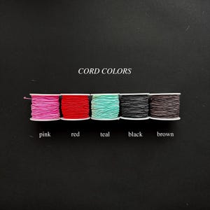 May include: Five spools of cord arranged in a horizontal row, featuring the colors pink, red, teal, black, and brown. The text CORD COLORS is centered above the spools, and the corresponding color names are printed below each individual spool.