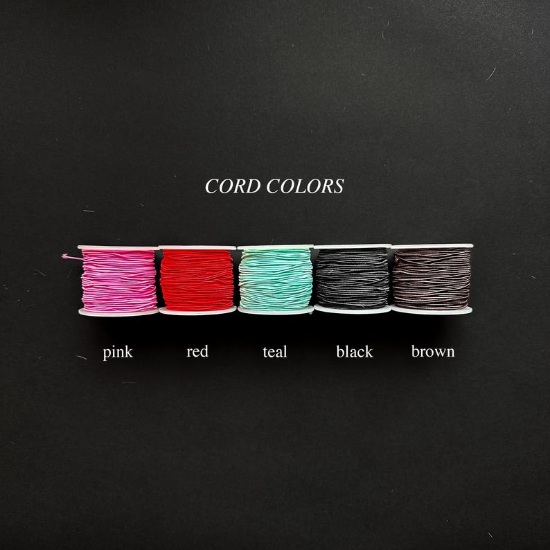 May include: Five spools of cord arranged in a horizontal row, featuring the colors pink, red, teal, black, and brown. The text CORD COLORS is centered above the spools, and the corresponding color names are printed below each individual spool.