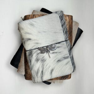 Puede incluir: Una pila de diarios de cuero con una variedad de patrones de piel de vaca en negro, marrón y blanco. El diario superior es blanco con marcas negras y un dije de libélula plateada. Una banda elástica negra asegura el diario.