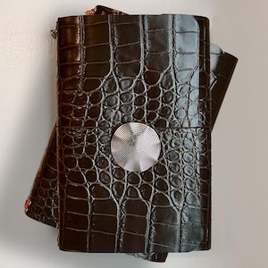 May include: A dark brown rectangular journal with a glossy crocodile skin texture. The cover features a centered, circular silver-toned metal embellishment with a fine, concentric spiral pattern etched into its surface, securing the journal closed.