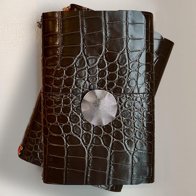 May include: A dark brown rectangular journal with a glossy crocodile skin texture. The cover features a centered, circular silver-toned metal embellishment with a fine, concentric spiral pattern etched into its surface, securing the journal closed.