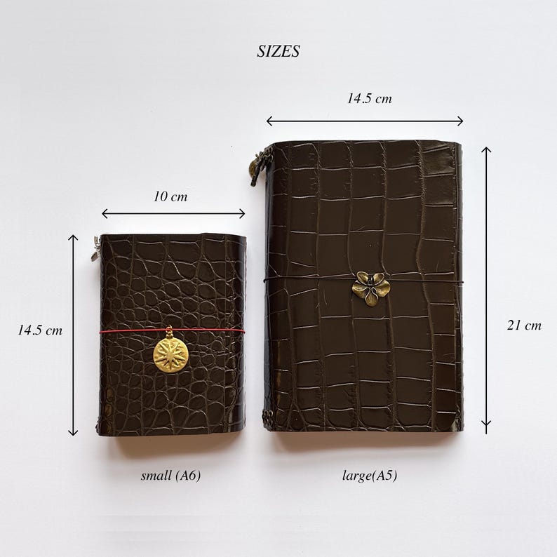May include: Two dark brown crocodile-textured journals with elastic closures. The small journal measures 10 cm by 14.5 cm and features a gold sun charm. The large journal measures 14.5 cm by 21 cm and features a gold flower charm. Text reads SIZES.