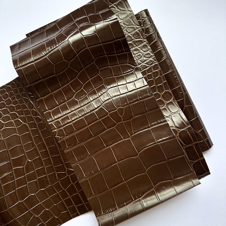 May include: A sheet of dark brown material featuring a glossy finish and a textured pattern that mimics the appearance of crocodile skin. The surface displays a repeating grid of irregular, raised rectangular and rounded scales in a deep brown.