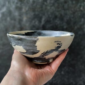 Peut inclure: Un bol en céramique présentant une base de couleur crème avec des motifs de coups de pinceau noirs audacieux et balayés sur l'extérieur. Le bol possède un bord large et ouvert ainsi qu'un petit pied circulaire surélevé à la base.