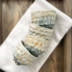 Pode incluir: Uma pilha de quatro pequenas tigelas de cerâmica com uma superfície texturizada e irregular. As tigelas apresentam um esmalte manchado em tons de bege, castanho e azul-acinzentado, com uma forma orgânica irregular e um rebordo espesso.