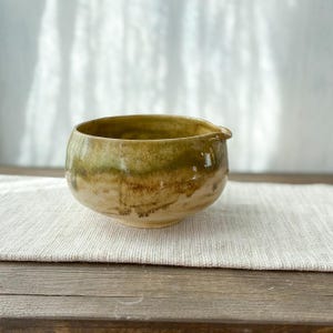 May include: A ceramic bowl with a rounded shape and a small pouring spout. The exterior features a variegated glaze with earthy tones of olive green, brown, and cream, showing a smooth, glossy finish with subtle variations in color intensity.