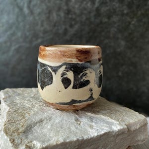 May include: A ceramic cup with a rounded shape, featuring a cream-colored base with bold, sweeping black brushstroke patterns around the center and a warm, earthy brown glaze around the top rim, showcasing a textured and hand-crafted appearance.