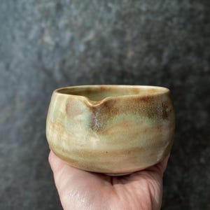 Może przedstawiać: Mała, okrągła ceramiczna miseczka z dzióbkiem do nalewania. Zewnętrzna strona pokryta jest zróżnicowaną glazurą w odcieniach beżu, jasnego brązu i delikatnej szałwiowej zieleni, co nadaje gładki, nakrapiany wygląd zakrzywionej powierzchni naczynia.