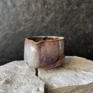 Op de afbeelding: Een kleine keramische creamer of schenkkan met een afgerond lichaam en een subtiele tuit. De buitenkant is voorzien van een reactief glazuur met gemengde tinten van gedempt paars, oudroze en zachtgrijs, wat zorgt voor een gladde, glanzende afwerking.