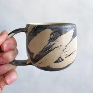 Pode incluir: Uma caneca de cerâmica com corpo arredondado e alça curva, apresentando um exterior cinza-carvão escuro com grandes padrões de pinceladas em um tom creme claro que revelam a textura da argila natural sob o esmalte escuro.