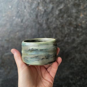 Op de afbeelding: Een kleine, ronde keramische beker met een gespikkeld glazuur in gemengde tinten saliegroen, leiblauw en antracietgrijs. Het oppervlak heeft een getextureerd, organisch uiterlijk met subtiele horizontale ribbels en een gladde, gebogen rand.