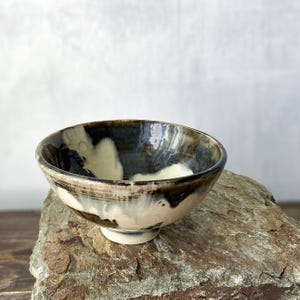 Może przedstawiać: Ceramiczna miska o zaokrąglonym kształcie i małej podstawie. Zewnętrzne i wewnętrzne powierzchnie prezentują marmurkowy wzór szkliwa z kontrastującymi wirami w kolorach głębokiej czerni, ciemnego brązu oraz kremowej bieli na całej powierzchni.