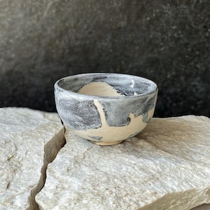 Pode incluir: Uma pequena tigela de cerâmica com um esmalte aplicado à mão num acabamento cinzento e branco manchado. As superfícies exterior e interior apresentam um padrão irregular e abstrato onde o esmalte cinzento escuro contrasta com a base de argila esbranquiçada.