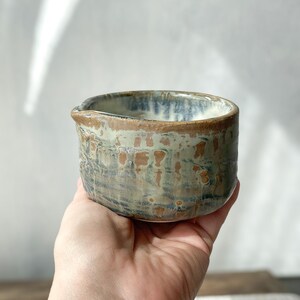Könnte beinhalten: Ein kleines Milchkännchen oder eine Gießschale aus Keramik mit einer reaktiven Glasur in gemischten Tönen aus erdigem Braun, gedämpftem Grün und sanftem Blau. Das Gefäß hat einen abgerundeten Körper, einen dicken Rand und einen kleinen Ausguss.