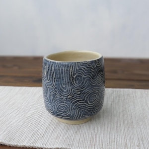 Może przedstawiać: Ceramiczny kubek o zaokrąglonym kształcie i gładkim rancie. Zewnętrzna strona pokryta jest ciemnoniebieskim szkliwem ze skomplikowanymi, jasnymi, wirującymi wzorami linii przypominającymi kontury mapy topograficznej. Wnętrze kubka jest kremowe.
