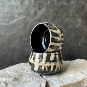 Puede incluir: Dos tazas de cerámica apiladas con un esmalte base negro oscuro y patrones irregulares de pinceladas verticales en color crema. El interior de la taza superior es negro liso y brillante, mientras que el exterior presenta un acabado texturizado.