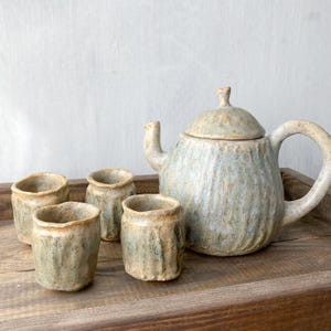 Può includere: Un set da tè in ceramica composto da una teiera con coperchio e tre tazze abbinate. I pezzi presentano una superficie testurizzata e scanalata con uno smalto maculato nei toni del blu tenue, verde salvia e beige terroso.