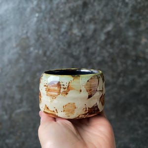 Pode incluir: Uma pequena chávena de cerâmica redonda com um esmalte de cor creme e manchas castanhas irregulares e mosqueadas na superfície exterior. O interior da chávena tem um acabamento em esmalte preto sólido, contrastando com o padrão exterior.