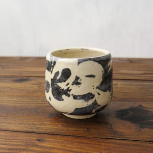 Może przedstawiać: Ceramiczny kubek o zaokrąglonym kształcie i szerokim otworze. Zewnętrzna strona ma kremową bazę z dużymi, abstrakcyjnymi wzorami w kolorze ciemnego antracytu, wykonanymi pociągnięciami pędzla, które owijają się wokół korpusu naczynia, tworząc kontrast.
