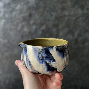 Op de afbeelding: Een kleine keramische schenkkom met een handmatig aangebracht glazuur in een gemarmerd patroon van diep marineblauw, crème en zwarte accenten. De binnenzijde heeft een gladde, olijfgroene afwerking en de rand is voorzien van een kleine schenktuit.