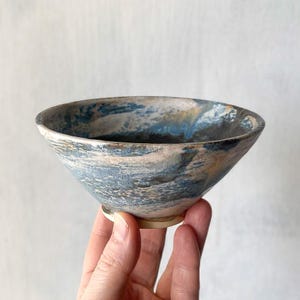 Può includere: Una ciotola in ceramica caratterizzata da uno smalto maculato nei toni del blu, bianco e marrone terra. La ciotola presenta un bordo ampio e aperto con una base affusolata, mostrando una superficie strutturata con visibili variazioni cromatiche.