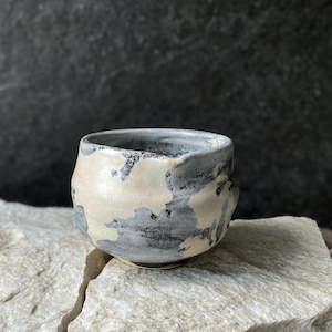 Op de afbeelding: Een kleine, ronde keramische beker met een gespikkeld, met de hand aangebracht glazuur in tinten crème, antracietgrijs en leiblauw. Het oppervlak heeft een onregelmatige, getextureerde afwerking met zichtbare kleurmenging en een brede, open rand.