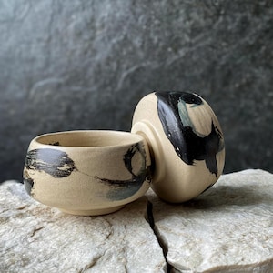 Può includere: Una ciotola da tè in ceramica con finitura beige opaca e audaci motivi a pennellate nere. La ciotola è accompagnata da un coperchio abbinato, appoggiato di lato, che mostra lo stesso design testurizzato dipinto a mano con inchiostro nero.